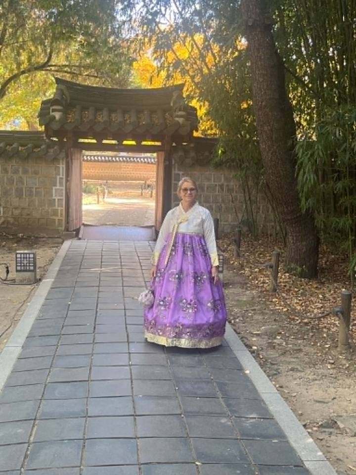 Woman wearing a purple hanbok stands in a traditional Korean courtyard surrounded by stone walls and bamboo.