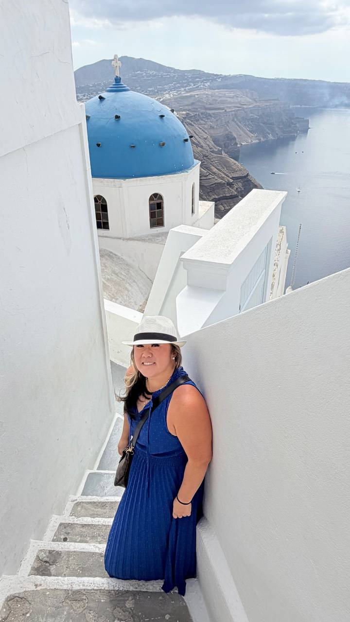 Close-up of a smiling woman in a white hat framed by stark white Santorini architecture and sea below.