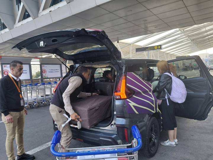 Travelers and driver load luggage into a van outside a modern airport terminal.