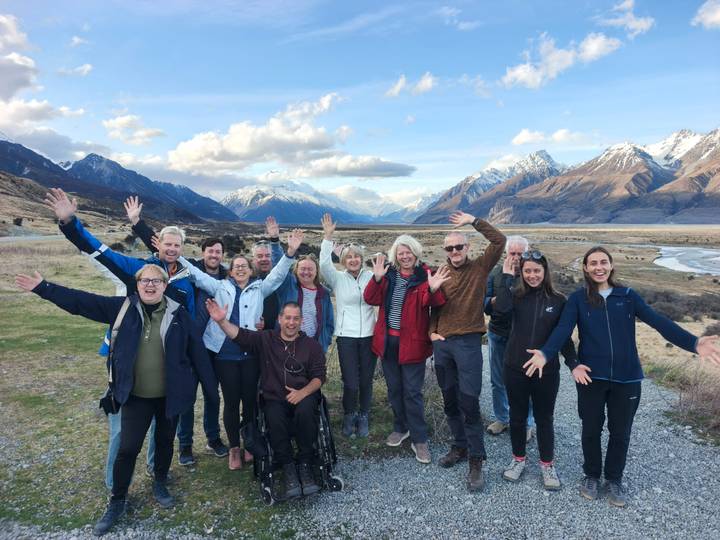 Smiling tour group raises hands with snow-capped Southern Alps and braided river behind.