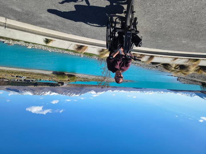 Man in wheelchair smiles with vivid turquoise Lake Tekapo and snowy peaks behind.