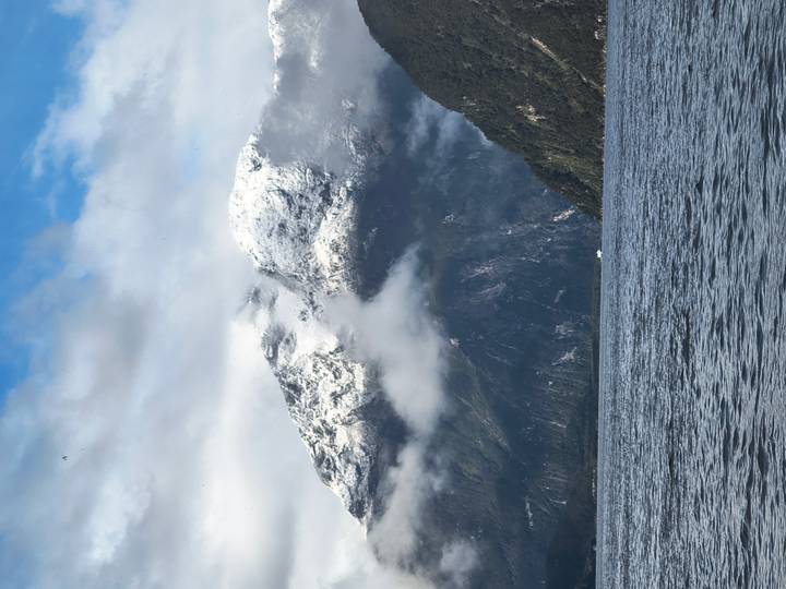 Snow-dusted peaks rise above misty fjord waters under patchy clouds.