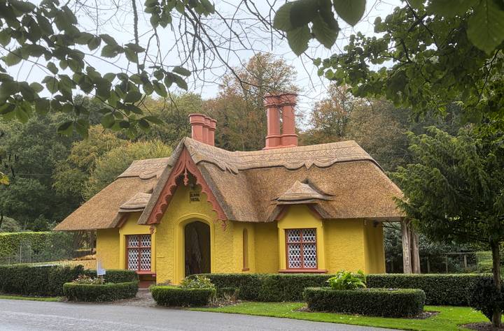 A quaint bright-yellow thatched cottage with ornate gables sits amid lush gardens and trees.