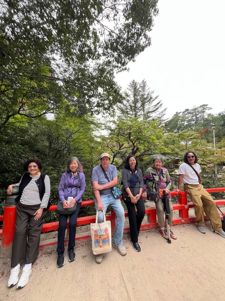 Travellers rest on a red wooden bridge in a leafy Japanese garden.