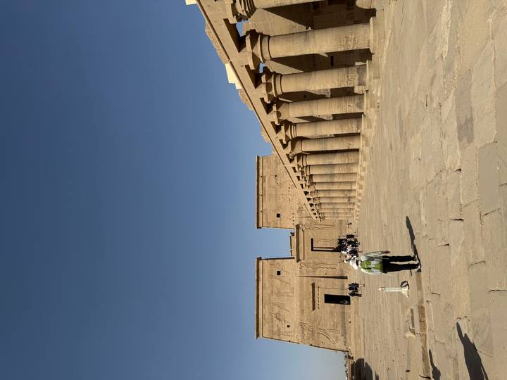 Visitors walk through a vast courtyard lined by tall stone columns and temple pylons under a deep blue sky