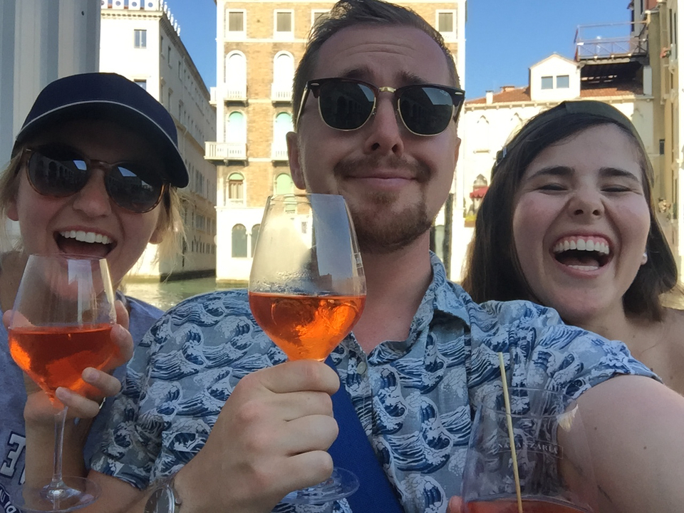 Three people smiling and holding glasses of drinks with a canal in the background.