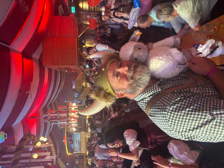 Man in Bavarian costume wearing horned helmet cuddles a plush sheep at a lively Oktoberfest beer hall.