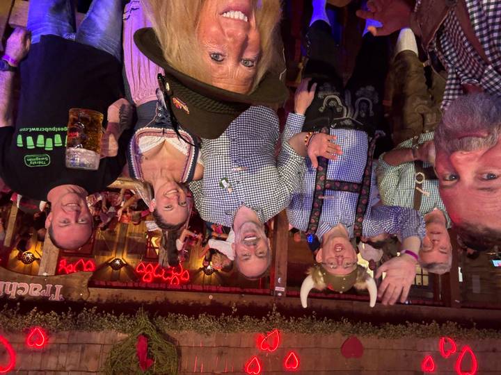 Smiling group in traditional Bavarian attire raise hands and beer steins inside a decorated beer tent.