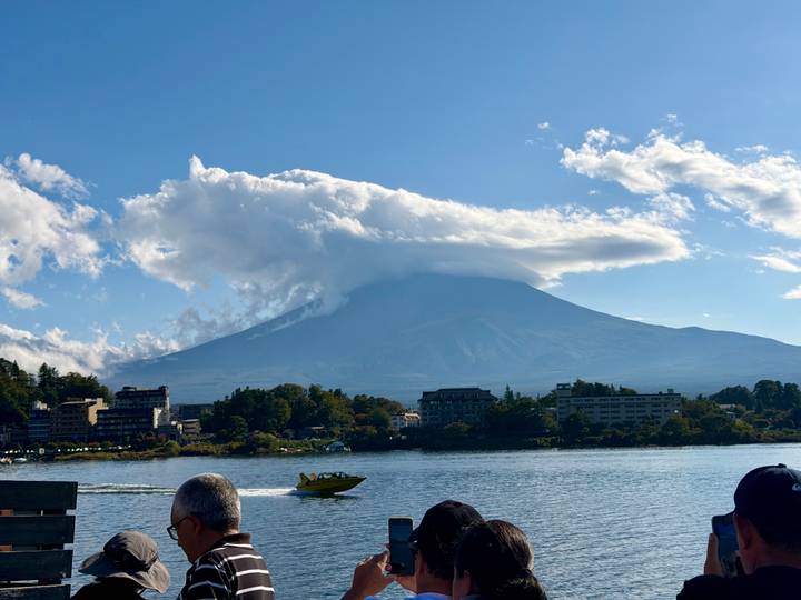 Mount Fuji capped by dramatic lenticular cloud looms above lakeside town and speeding yellow boat.