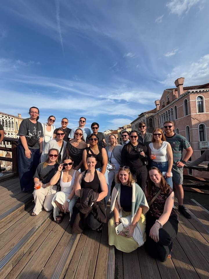 Large travel group poses on a Venetian wooden bridge with historic pastel buildings behind them.