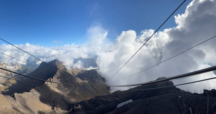 Sweeping alpine panorama with rugged peaks, clouds rolling through valleys and cable lines in view.