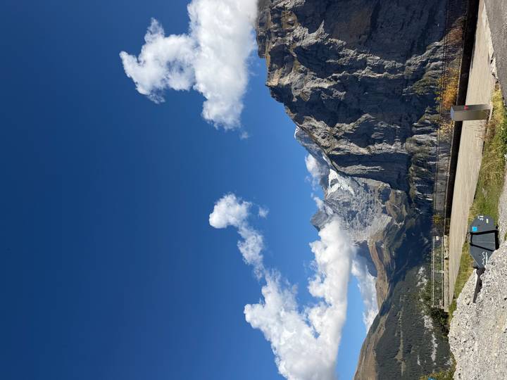 Snow-dusted Alpine peaks rise sharply under a vivid blue sky above a valley viewpoint.