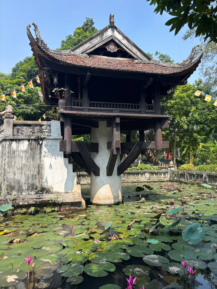 Traditional One-Pillar Pagoda standing over a lotus pond surrounded by greenery and prayer flags