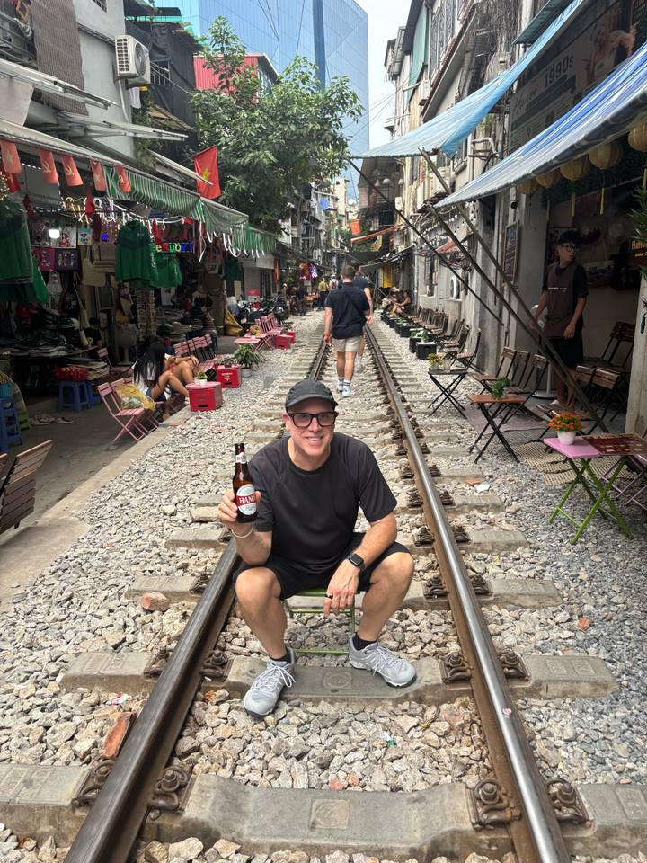 Tourists relaxing on fold-out chairs along Hanoi Train Street while a man poses with a beer on the rails