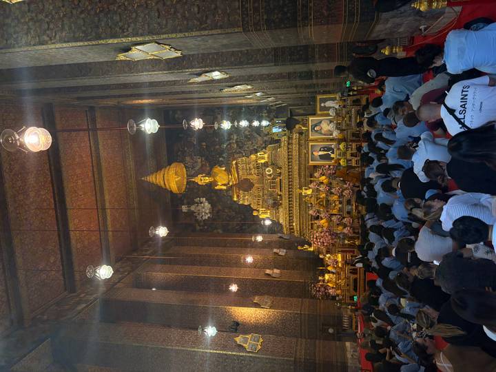 Interior of a grand Thai temple with a towering golden Buddha statue as worshippers sit on the floor in prayer.