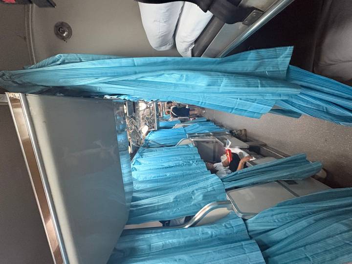 Narrow aisle of an overnight train with blue privacy curtains and passengers settling into bunk beds.