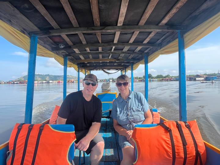 Smiling couple seated on a narrow longtail boat cruising across a vast lake with floating homes.