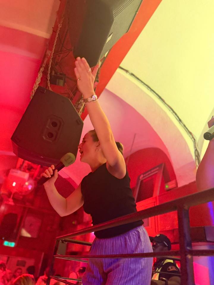 Woman passionately singing into a microphone under vibrant red club lighting.