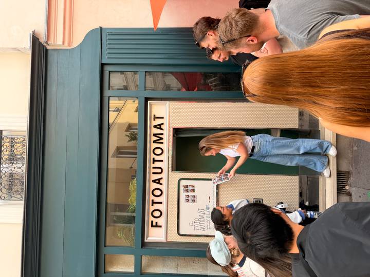 Crowd gathers around a retro street photo booth as a woman examines freshly printed strips.