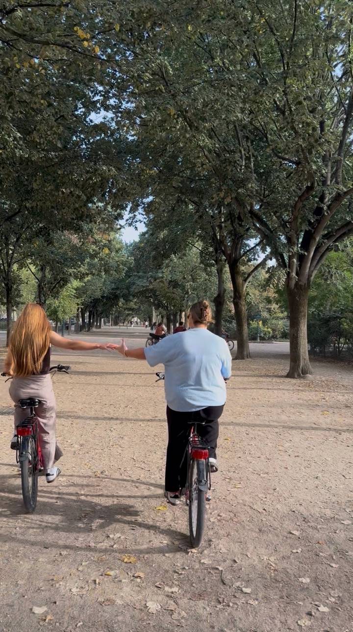 Two young women riding bicycles on a tree-lined dirt path, reaching out to hold hands under dappled sunlight.