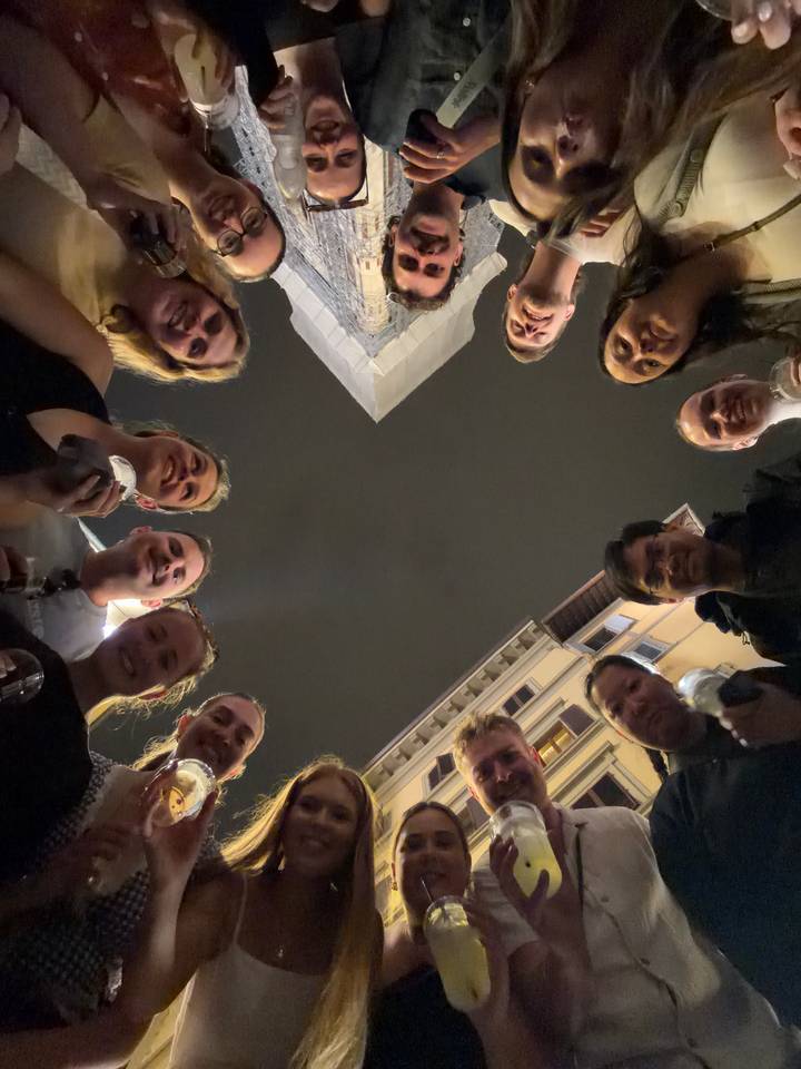 Night-time selfie taken from below with a circle of smiling friends holding drinks, open sky and surrounding buildings visible.