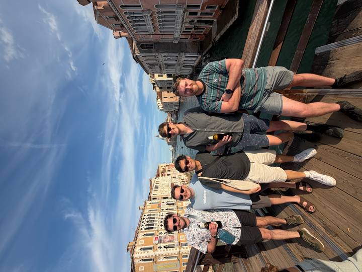Five friends posing on a wooden bridge over Venice's Grand Canal with historic palaces and blue sky.