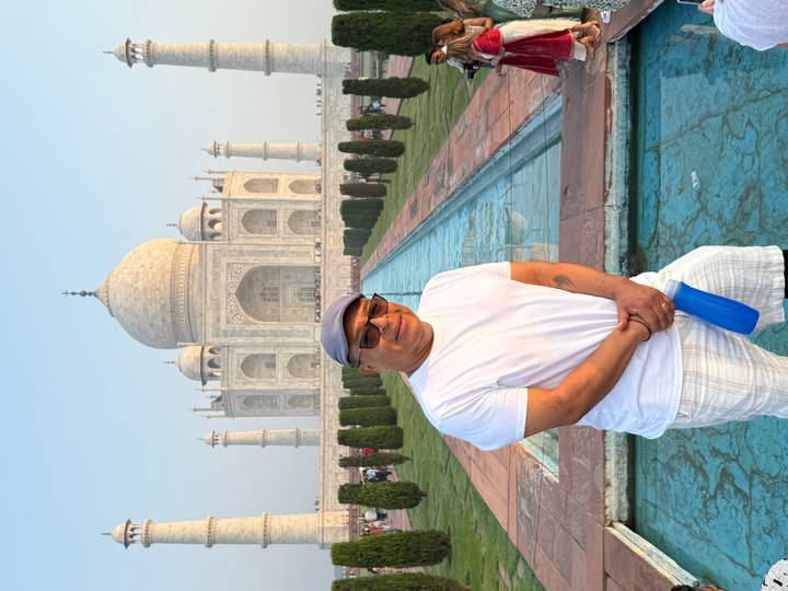 A man poses in front of the reflecting pool leading to the Taj Mahal under a soft morning sky.