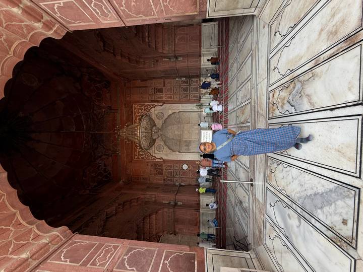 Visitors sit and walk inside a vast red-sandstone mosque hall while a woman in a sari stands for a photo.