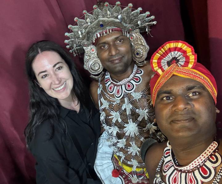 A smiling visitor takes a selfie with two Sri Lankan traditional dancers wearing ornate beadwork and headgear.