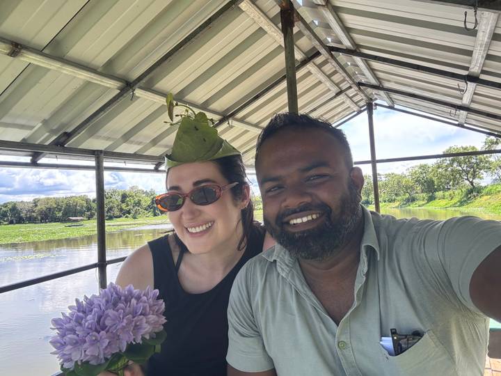 Two friends smile for a selfie on an open boat gliding across a calm lake under a leafy roof.