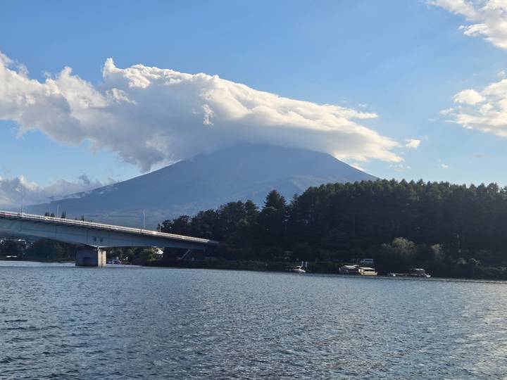 Clouds cloak the summit of Mount Fuji rising above a tranquil lake crossed by a modern bridge.