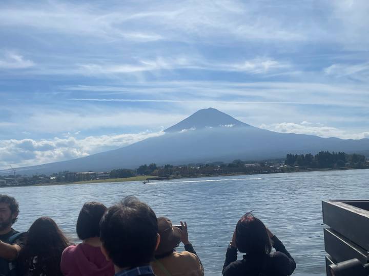 Mount Fuji rises above a lakeside town while tourists view the scene from a boat.