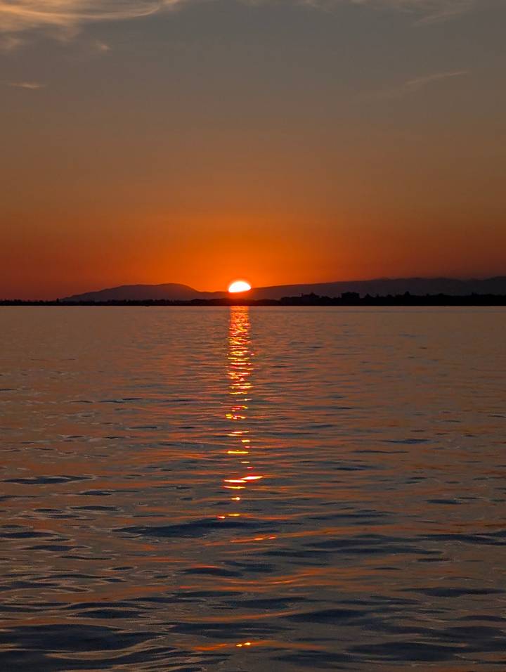 Serene sunset with the sun dipping behind distant mountains and its reflection stretching over a lake.