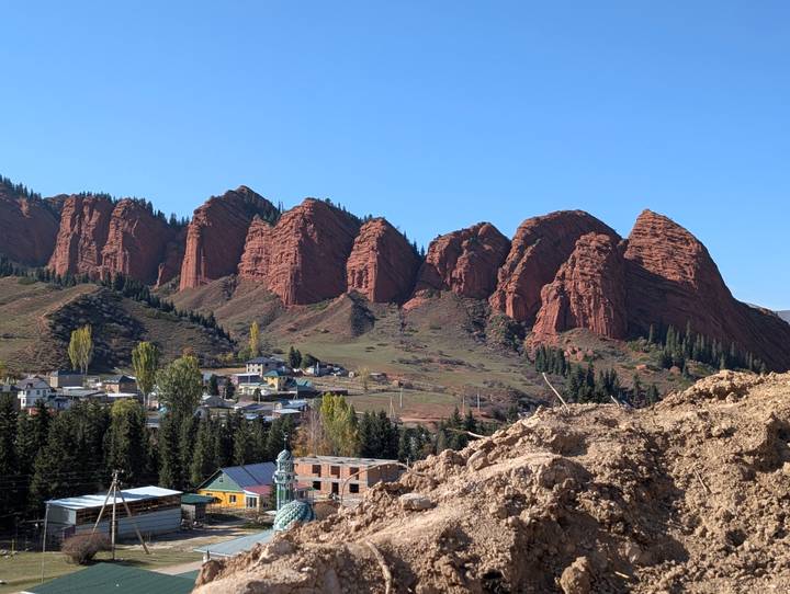 Red sandstone cliffs of Jeti-Oguz rising behind a small Kyrgyz village and green valley.
