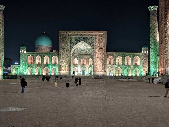 Registan square in Samarkand illuminated in green and gold lights at night with visitors walking across the plaza.