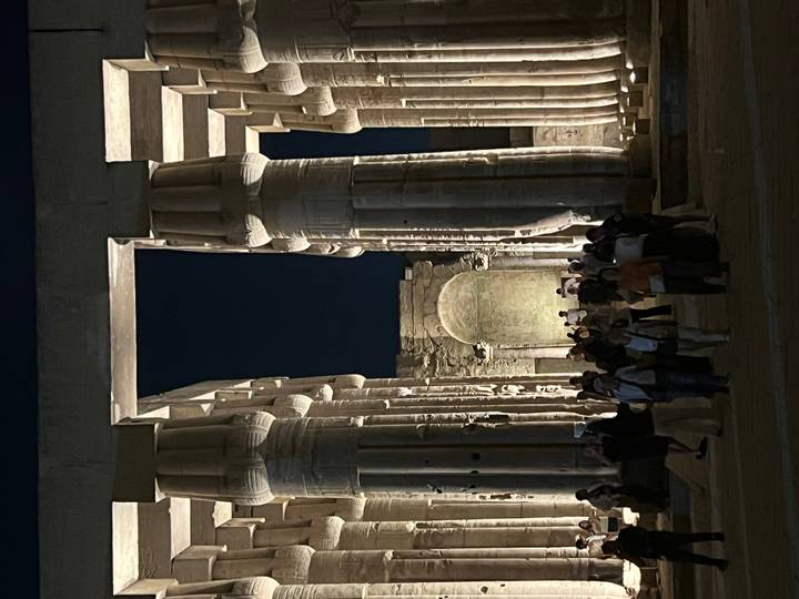 Nighttime visitors stand between towering lit columns of Kom Ombo Temple against a dark sky.