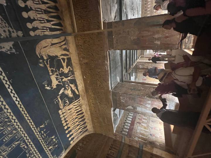 Interior of ancient tomb with painted ceiling and walls; tourists exploring in low light.
