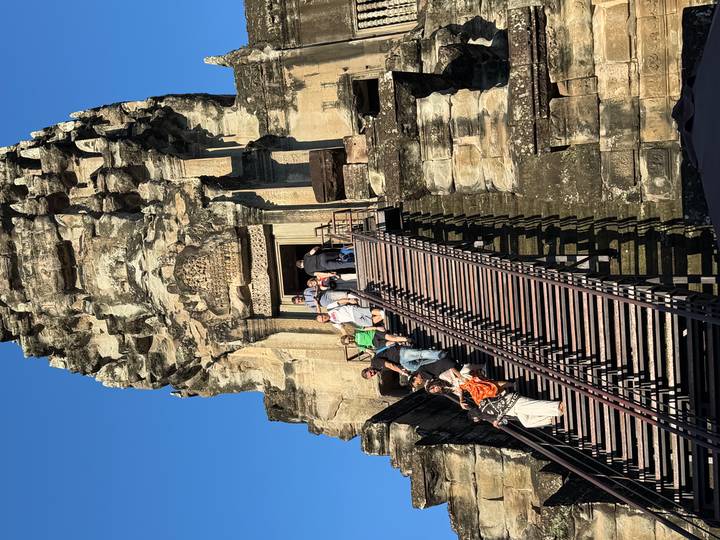Tourists climb the steep wooden stairs of an ancient Angkor temple tower under a clear blue sky.