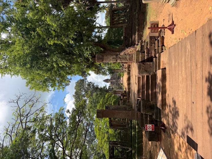 A wooden walkway leads through columned ruins toward a distant Angkor tower framed by leafy trees.