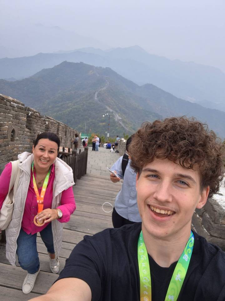 Selfie with two travellers smiling on the Great Wall with mountain ridges behind and other visitors on the walkway.