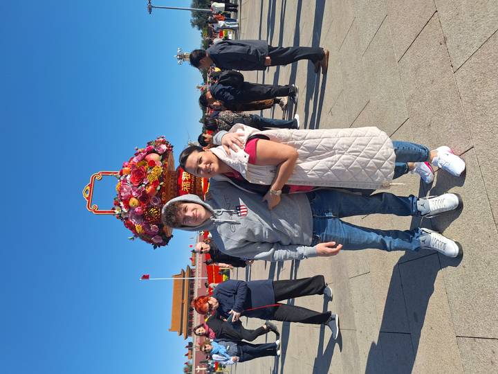Mother and son smiling in Tiananmen Square under clear blue sky with giant flower basket decoration behind.