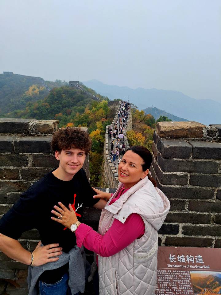 Two travellers posing on a tower of the Great Wall with autumn foliage and a long line of visitors stretching into the hills.