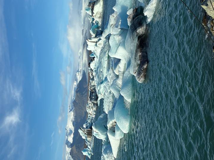 Expansive view of turquoise water dotted with many pale blue icebergs and snow-capped mountains behind.