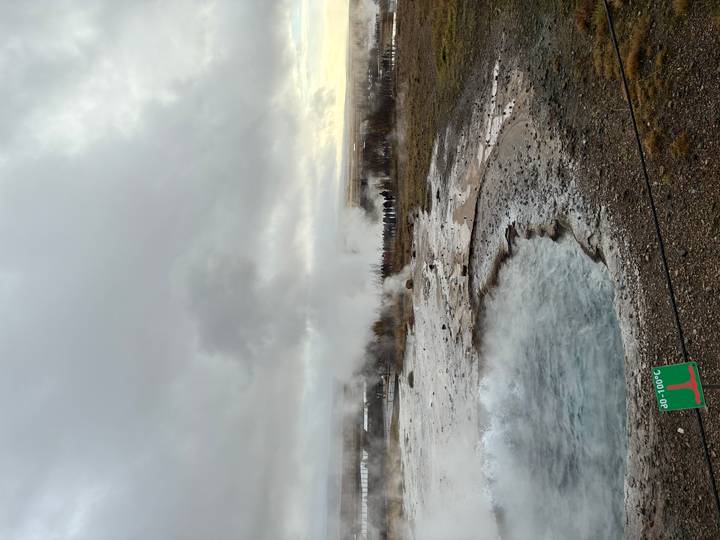 Steaming geothermal pool and erupting geyser plume rising into a cloudy sky.