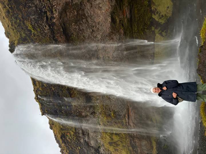 Elderly traveler standing in front of a tall, narrow waterfall cascading from a mossy cliff.