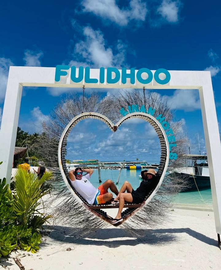Two men relax on a heart-shaped swing beneath a Fulidhoo island sign with turquoise water behind.