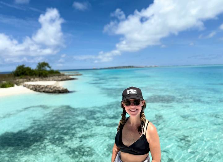 Smiling woman in a cap stands before crystal-clear turquoise lagoon and white sand island.