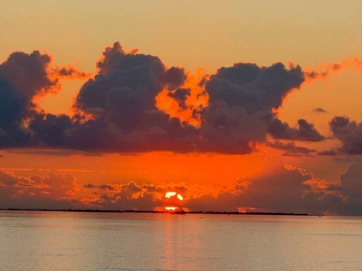 Vibrant orange sunset dipping below a low cloud bank over calm sea waters.
