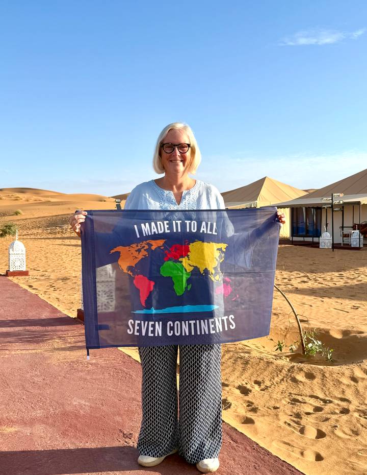 Smiling traveller in a desert camp holds a flag proclaiming she has visited all seven continents.