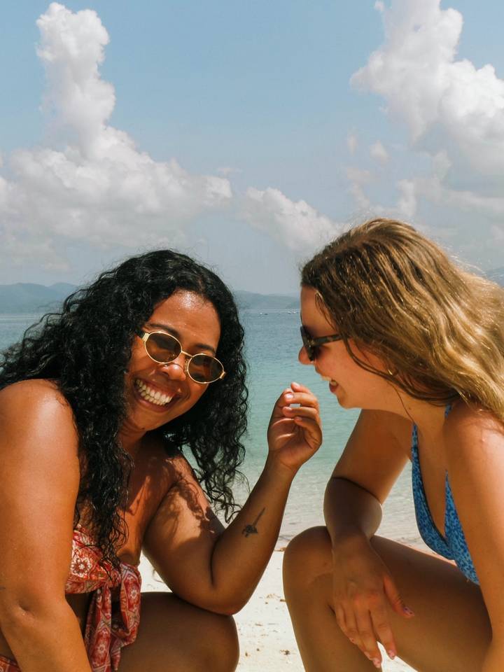 Two female friends laugh together on a tropical beach with turquoise sea and hazy mountains beyond.